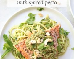 This is an overhead image of salmon with pesto zoodles on a white plate. The dish is garnished with cashews and cilantro. The plate sits on a white counter. Gold forks are on the counter next to the plate to the right. Text overlay reads "salmon + zoodles with spiced pesto."
