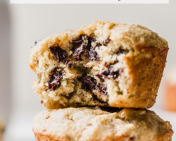 This is a side view of two banana muffins stacked on top of each other. The top muffin has a bite taken out revealing melty chocolate chips. The muffins sit on a white surface with a banana blurred in the background. Text overlay reads "chocolate chip banana muffins made with almond flour."