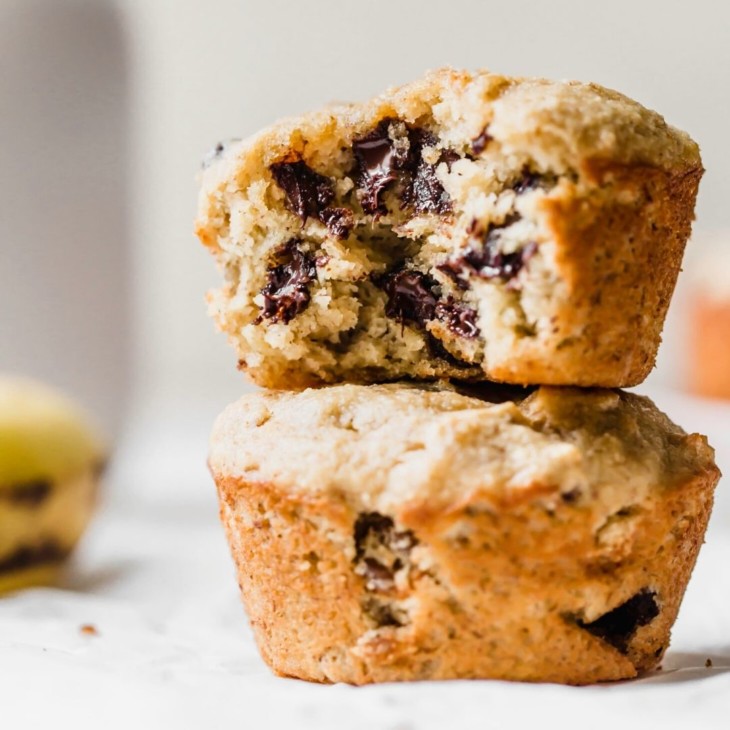 This is a side view of two banana muffins stacked on top of each other. The top muffin has a bite taken out revealing melty chocolate chips. The muffins sit on a white surface with a banana blurred in the background.