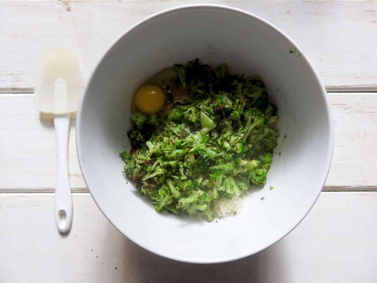 This is an overhead image of a white bowl with broccoli florets, an egg, and some shredded cheese. The bowl sits on white wooden planks and a white spatula lays next to the bowl.