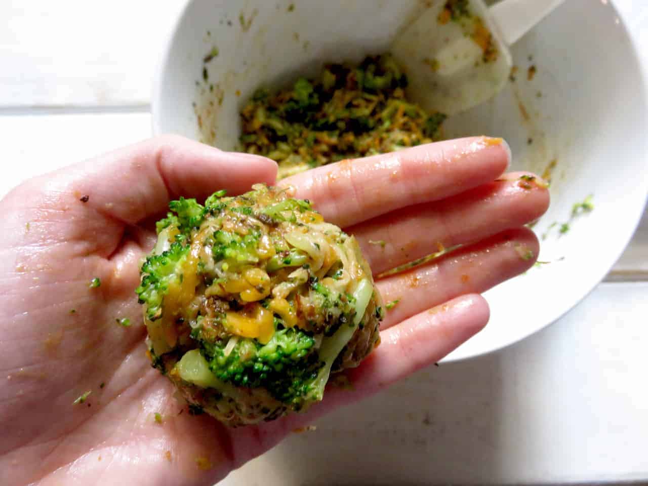This is a hand holding a broccoli cheddar mixture that was formed into a small patty. A white bowl is in the background with more broccoli cheddar mixture. 