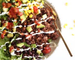 This is an overhead image of a bowl filled with lettuce, ground beef, salsa, avocado, corn, black beans, and sour cream. The bowl sits on a white counter and a gold fork lies partially in the bowl.