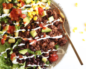 This is an overhead image of a bowl filled with lettuce, ground beef, salsa, avocado, corn, black beans, and sour cream. The bowl sits on a white counter and a gold fork lies partially in the bowl.
