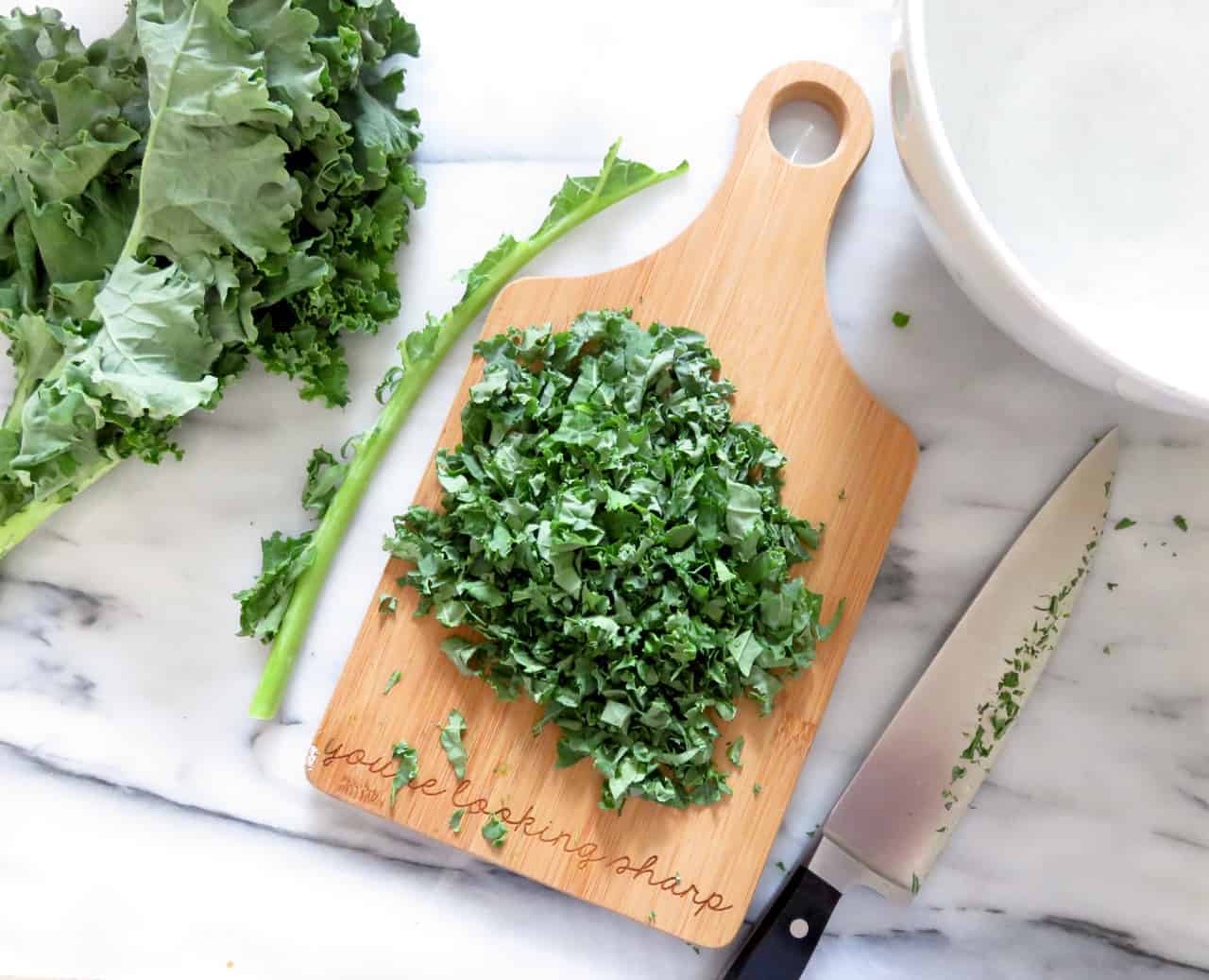 this is an overhead image of a cutting board with chopped kale on it. the cutting board sits on a white marble counter. To the left of the cutting board is more kale and a stem of kale. to the right top corner of the image is a large white bowl. 