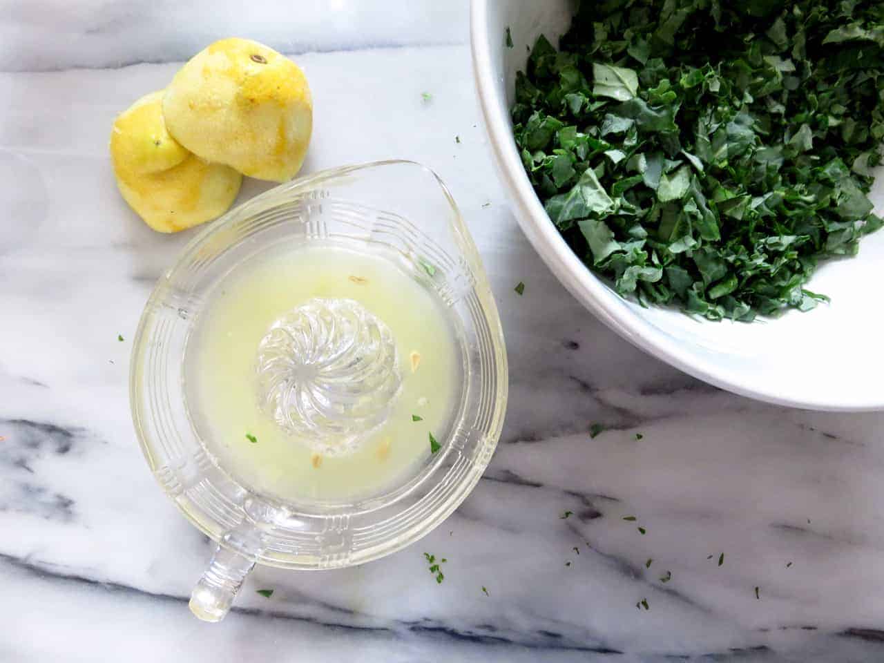 this is an overhead image of a large white bowl with chopped kale. a juicer has lemon juice in it with lemon halves above the juicer on the white marble counter.