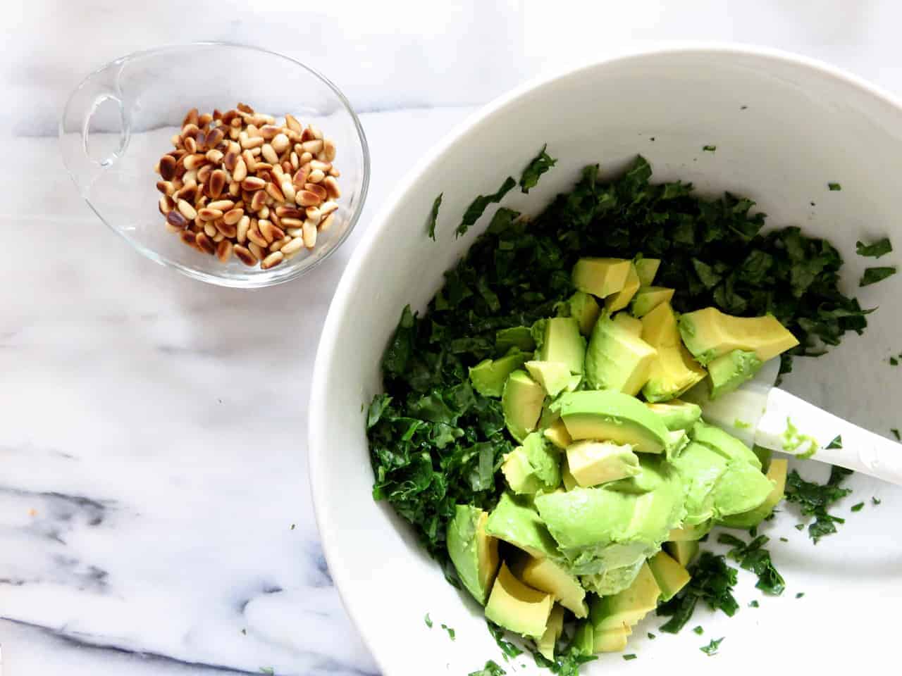 this is an overhead image of a white bowl with chopped kale and avocado chunks. The bowl sits on a white marble counter with a small glass bowl of toasted pine nuts.