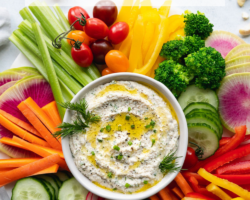 This is an overhead image of a white bowl filled with a creamy dip. The dip is topped with a drizzle of olive oil and fresh herbs. Around the dip are fresh veggies like cut peppers, radish, carrots, cucumbers, and celery. Text overlay reads "Vegan Ranch Dip made with cashews!"