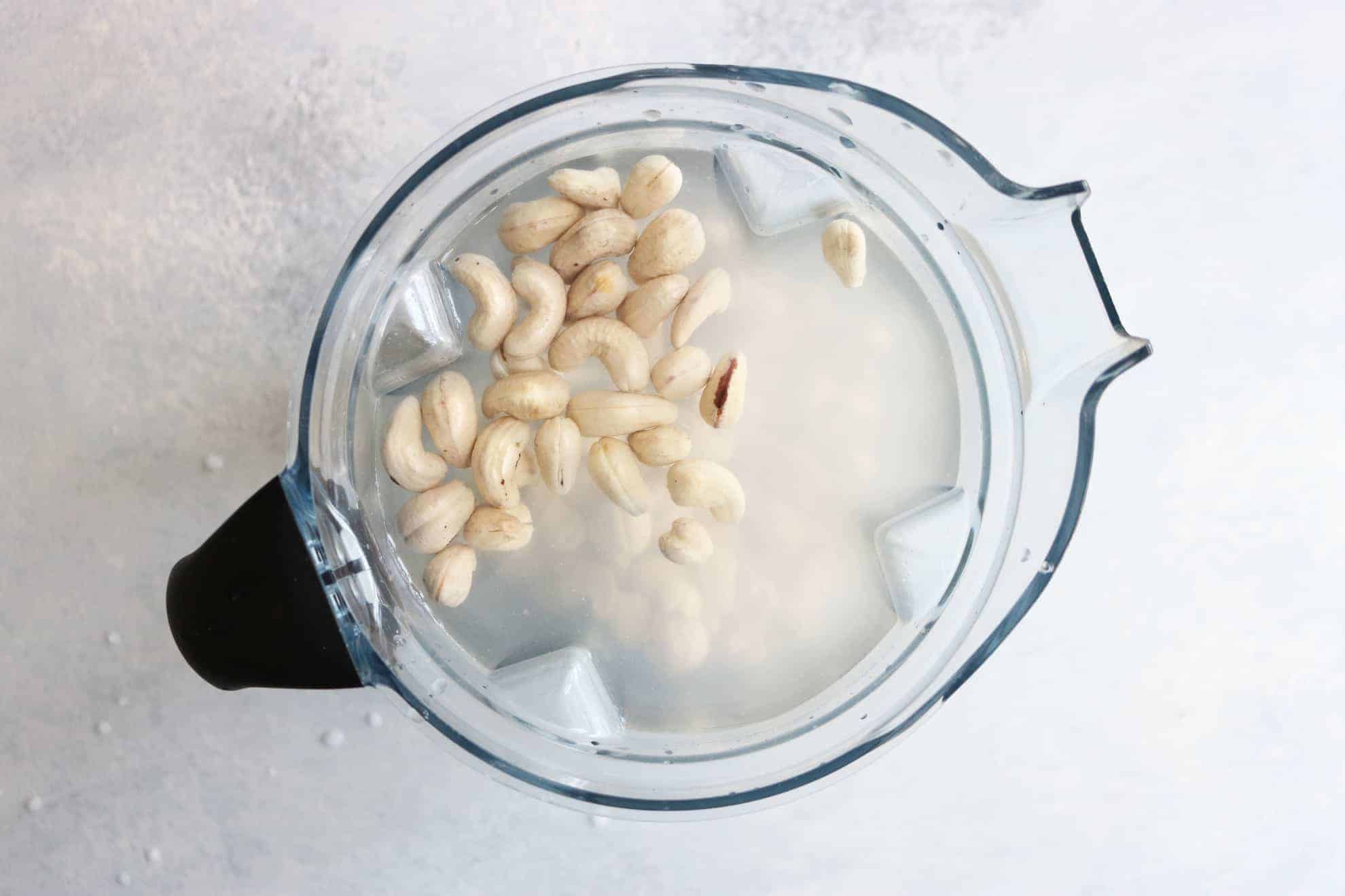This is an overhead image of a blender filled with water and cashews. The blender sits on a white and grey surface. 