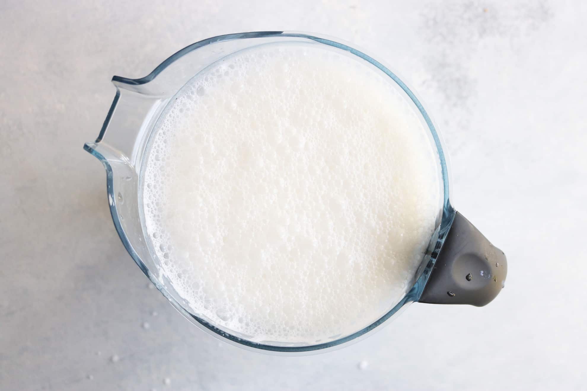 This is an overhead image of a blender with blended milk. The milk has a lot of bubbles. The blender sits on a light grey surface.