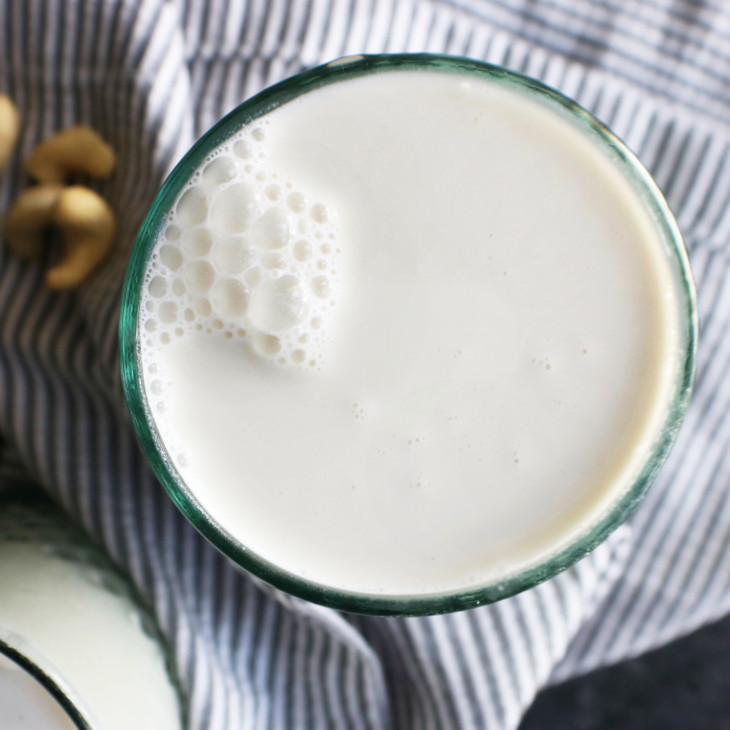This is an overhead image of a glass filled with milk. The glass sits on a grey pinstripe tea towel on a dark grey surface. Cashews are scattered around the glass.