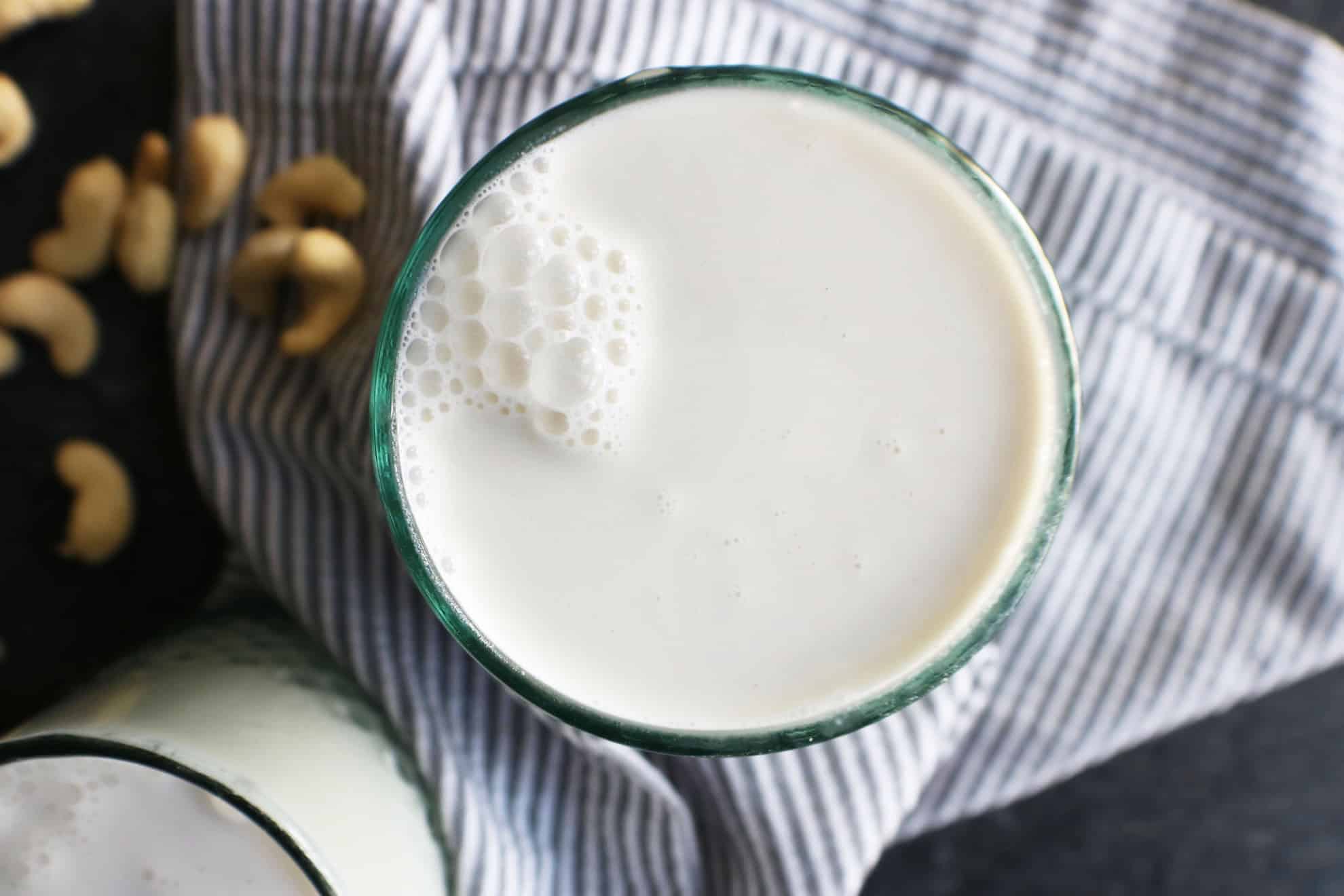 This is an overhead image of a glass filled with milk. The glass sits on a grey pinstripe tea towel on a dark grey surface. Cashews are scattered around the glass. 