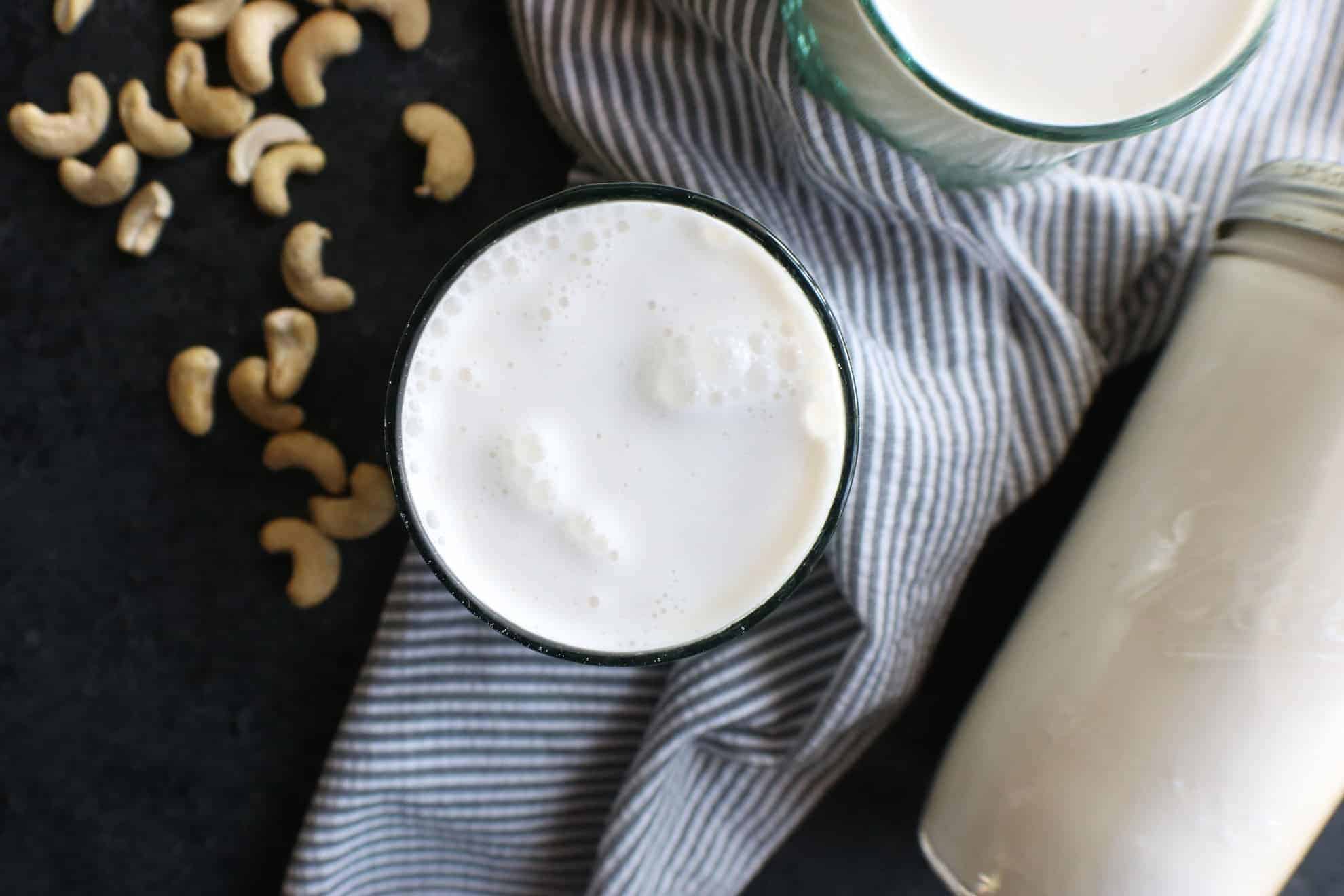 This is an overhead image of a glass filled with milk. The glass sits on a grey pinstripe tea towel on a dark grey surface. Cashews are scattered around the glass. A mason jar filled with milk is off to the bottom right of the image.