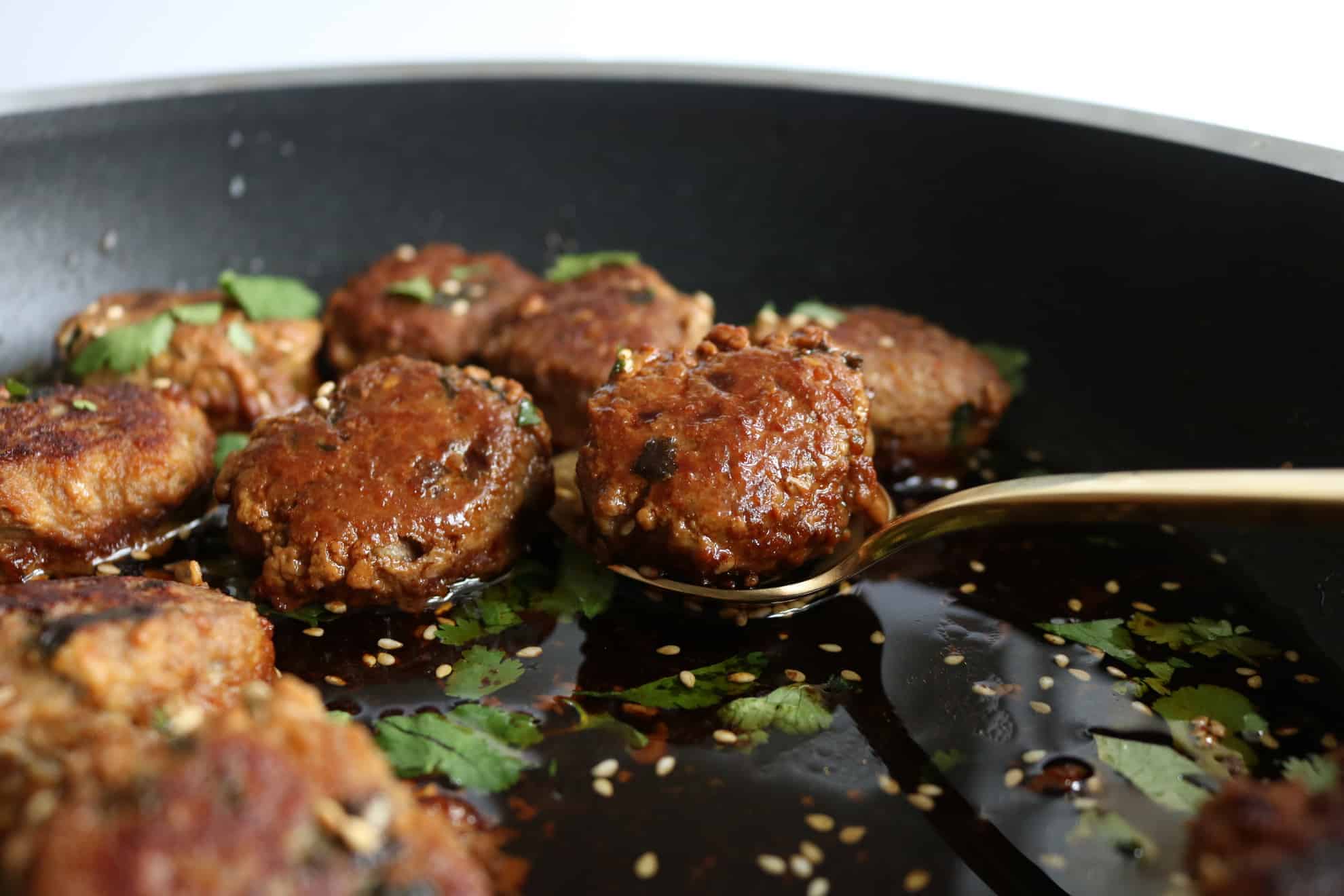 This is a side view of turkey meatballs in a pan. A gold spoon is scooping up the meatball. The meatballs are in a soy sauce with sesame seeds and cilantro. 