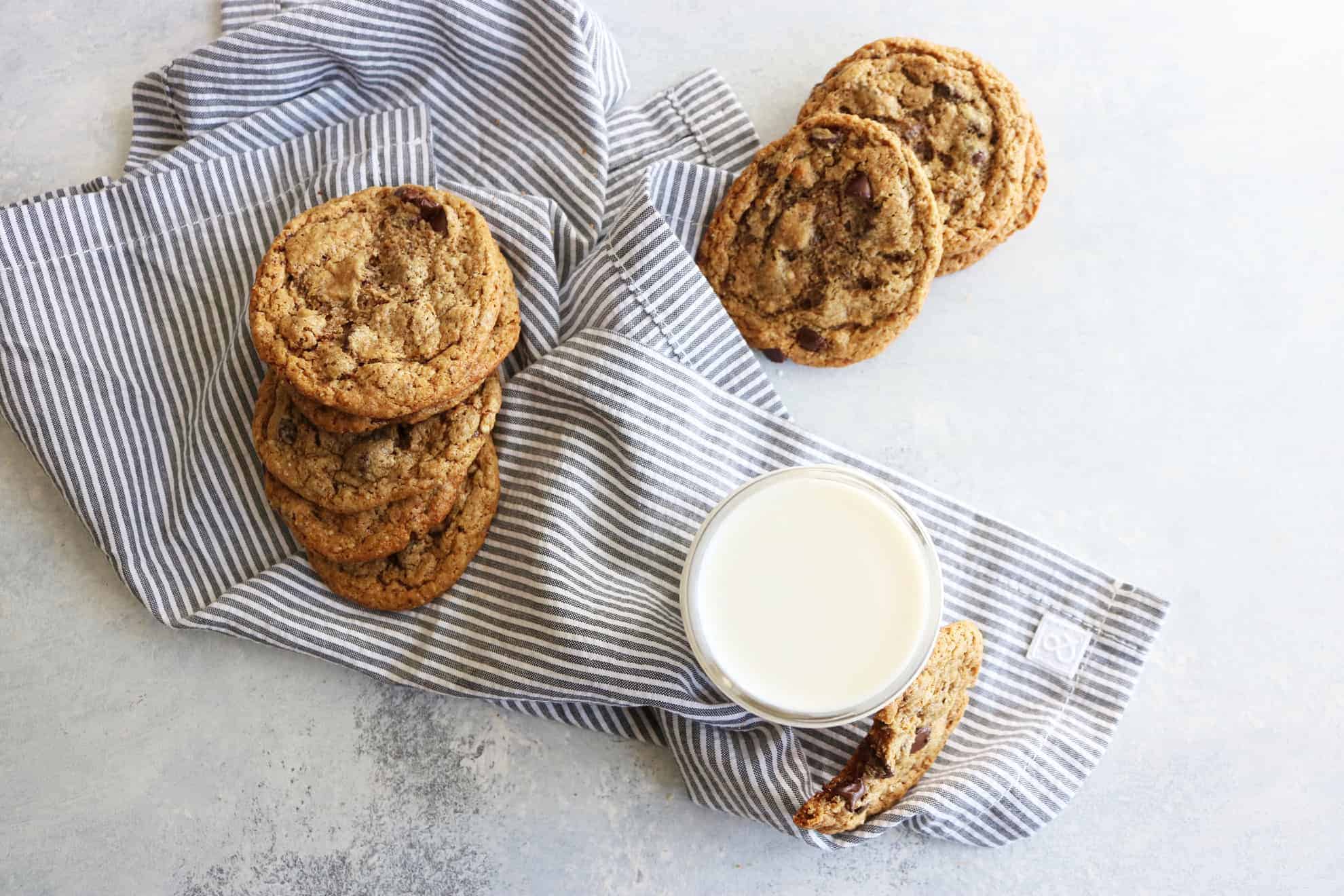 This is an overhead image of a glass of milk with a cookie leaning against it. The cookie has a bite taken out. More cookies are off to the side and around the glass of milk. The glass sits on a white and grey surface with a grey pinstripe tea towel. 