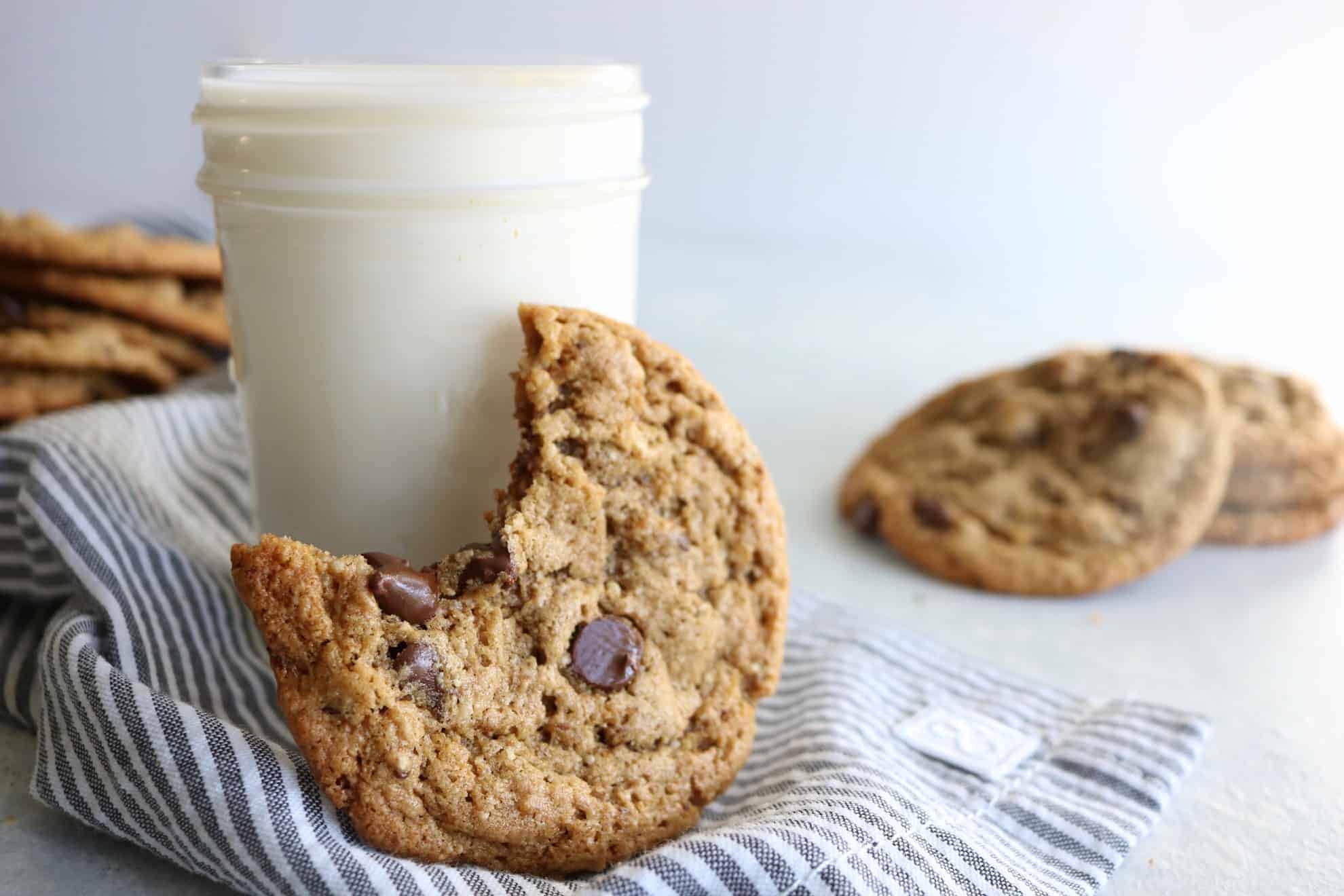 This is a side image of a glass of milk with a cookie leaning against it. The cookie has a bite taken out. More cookies are off to the side and blurred in the background. The glass sits on a white and grey surface with a grey pinstripe tea towel. 