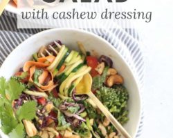 This is an overhead image of zoodle salad. A gold fork is twirling zoodles around the fork. The salad bowl sits on a whitw counter with grey pinstripe tea towel. Text overlay reads "zoodle salad with cashew dressing."