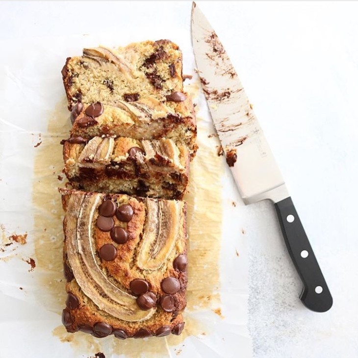 This is an overhead image of a banana bread with sliced banana on top and chocolate chips. A sharp knife lays next to the bread. The bread sits on a white piece of parchment paper.
