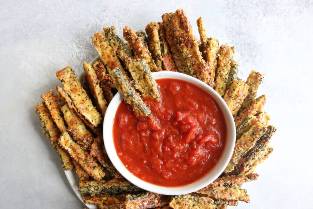 This is an overhead image of a plate with parmesan rusted zucchini fries on a plate. The plate sits on a white counter. The fries are dipped in a smaller bowl of marinara sauce.