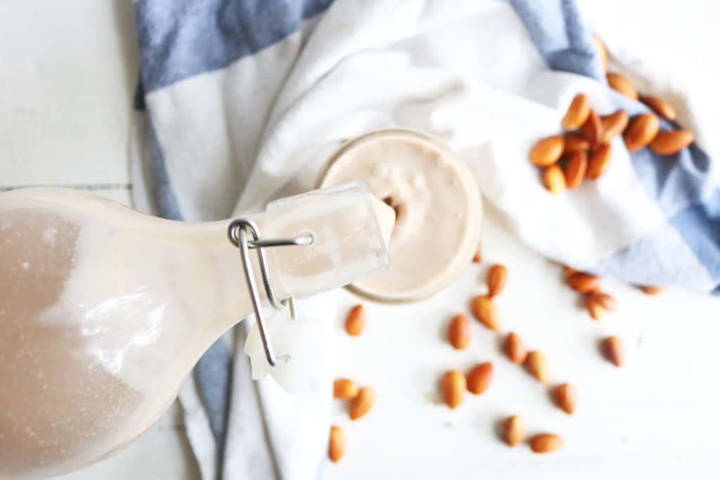 This is an overhead image of a glass pitcher pouring milk into a glass. The glass is sitting on a white surface with almonds and a white and blue tea towel. 