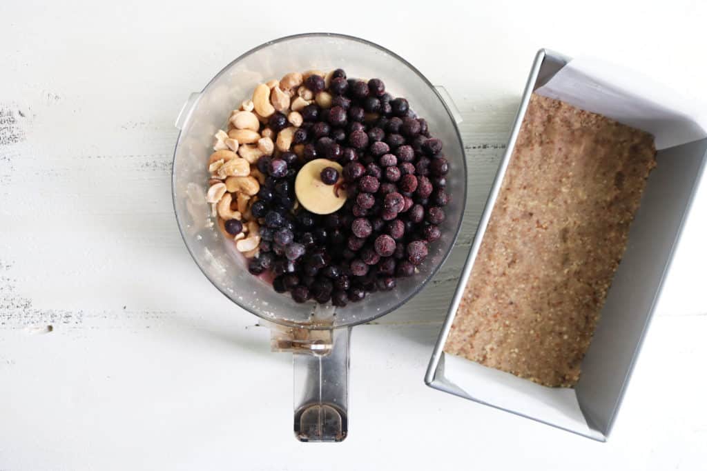 This is an overhead horizontal image of a food processor with pecans and frozen blueberries in it. To the right of the food processor is a bread pan lined with parchment paper and a nutty crust pressed into the bottom of it. The food processor and pan sit on a white wood surface. 