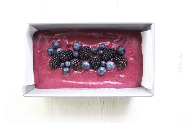 This is an overhead horizontal image of a silver bread pan lined with parchment paper. In the bread pan is a deep purple mixture topped with blueberries and blackberries. The pan sits on a white surface. 