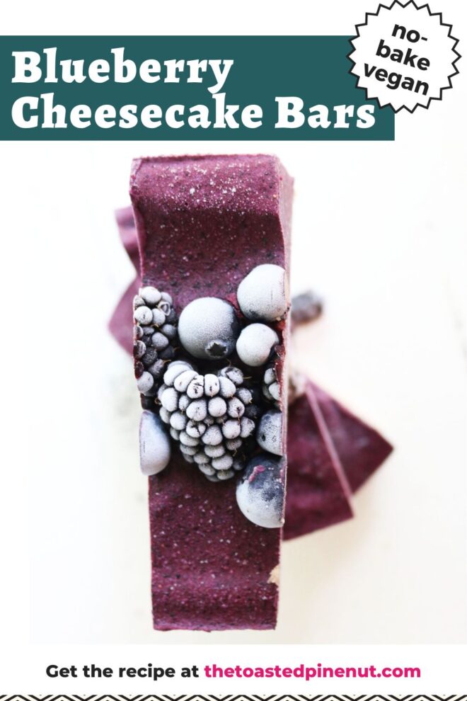 This is an overhead vertical image of a deep purple bar stacked on other bars beneath it. The bar has frozen, frosted blueberries and blackberries on top of it. The bars sit on a white surface. Text overlay reads "blueberry cheesecake bars no-bake vegan"