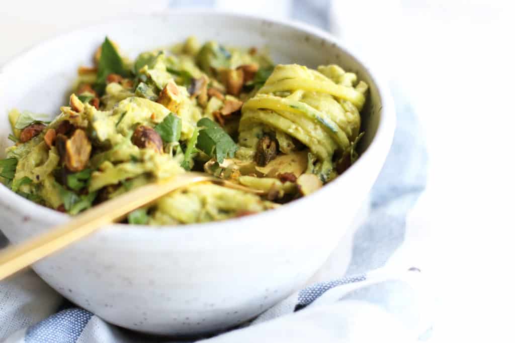 This is a side view of a small white bowl while with zoodles. A fork twirls the zoodles around the fork and leans against the side of the bowl. The bowl is topped with pistachios. 