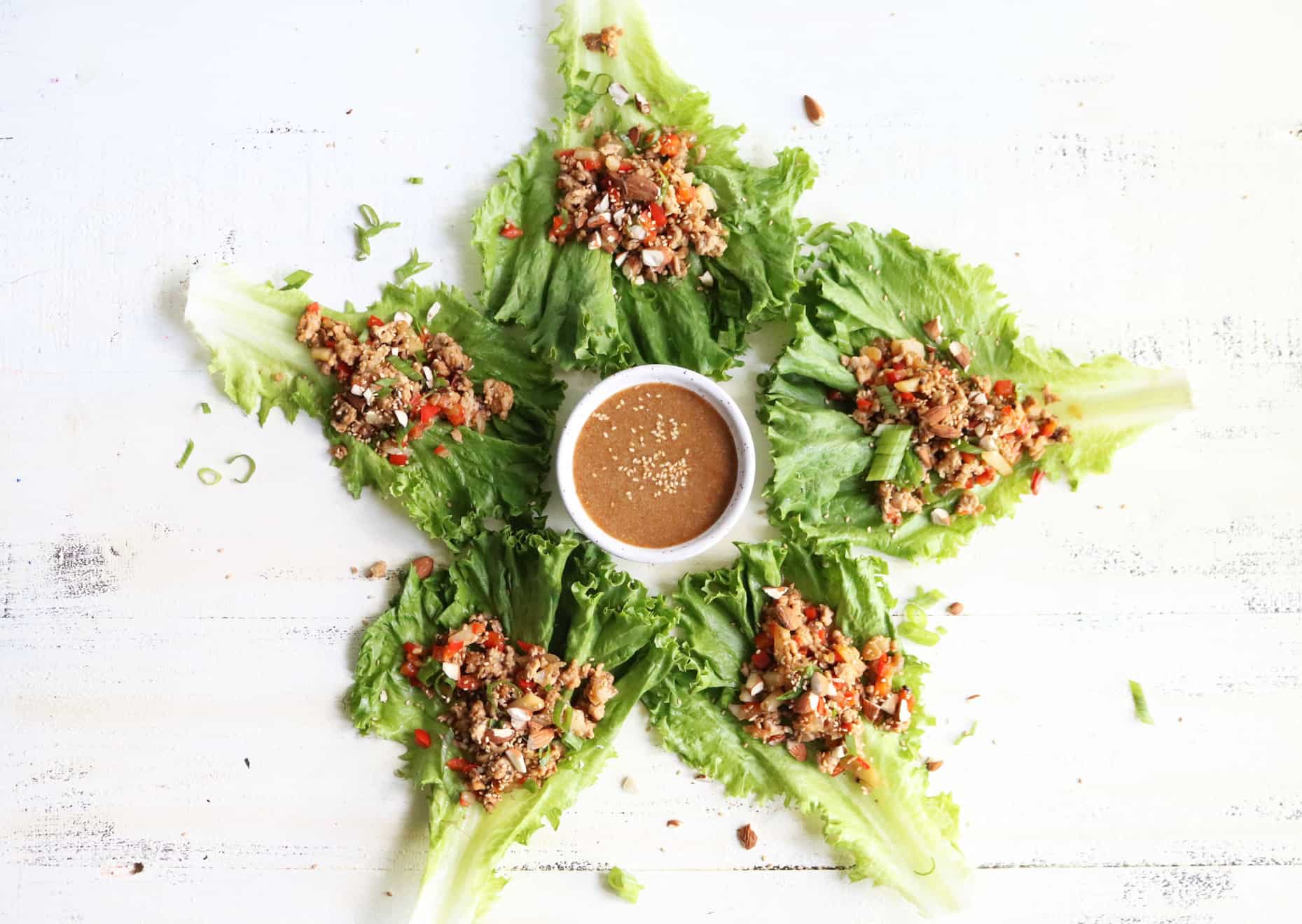 This is an overhead view of five chicken lettuce wraps on a white surface. The lettuce wraps are arranged around a small white bowl with sauce in it. 