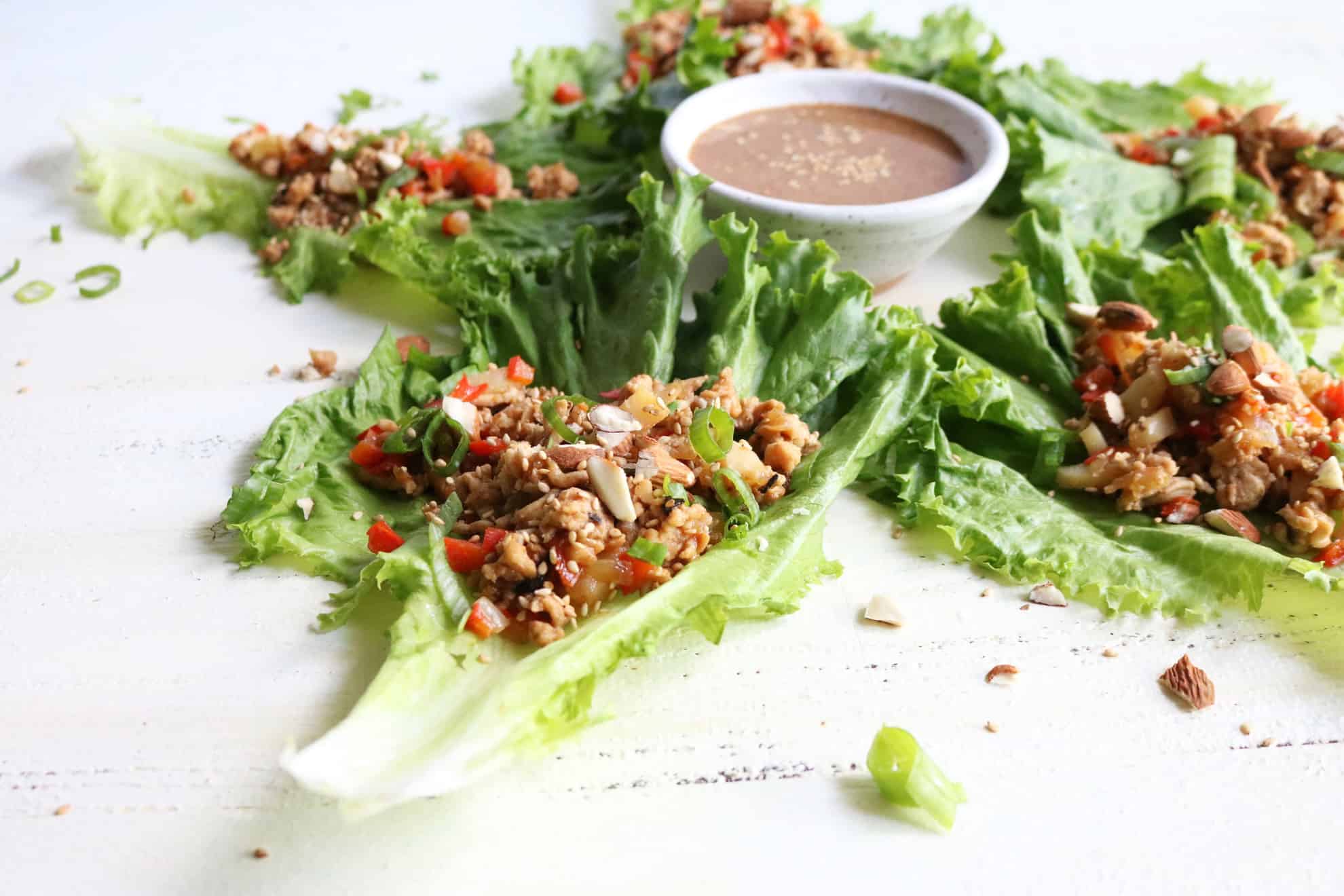 This is a side view of chicken lettuce wraps on a white surface. The lettuce wraps are arranged around a small white bowl with a sauce in it. 
