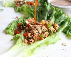 this is a side view of a chicken lettuce wrap with sauce being pour on top. The lettuce wrap sit on a white surface with more lettuce wraps blurred in the background.