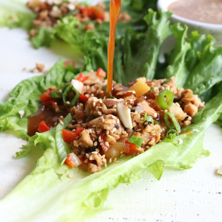 this is a side view of a chicken lettuce wrap with sauce being pour on top. The lettuce wrap sit on a white surface with more lettuce wraps blurred in the background.