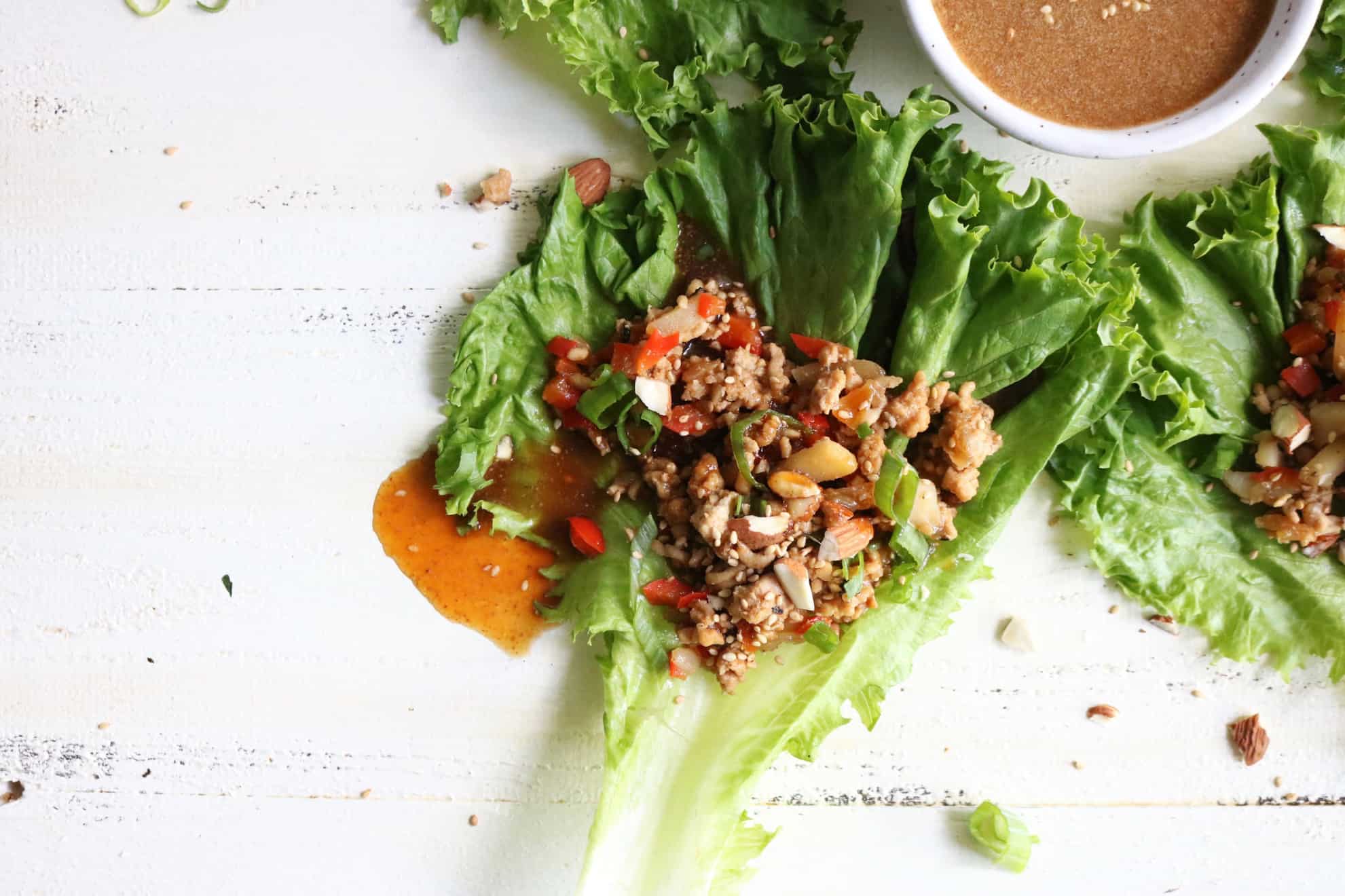 This is an overhead image of a chicken lettuce wrap with sauce dripping out the side. The lettuce wrap is on a white surface with other lettuce wraps to the right side and upper part of the image. A small sauce bowl is in the top right of the image. 