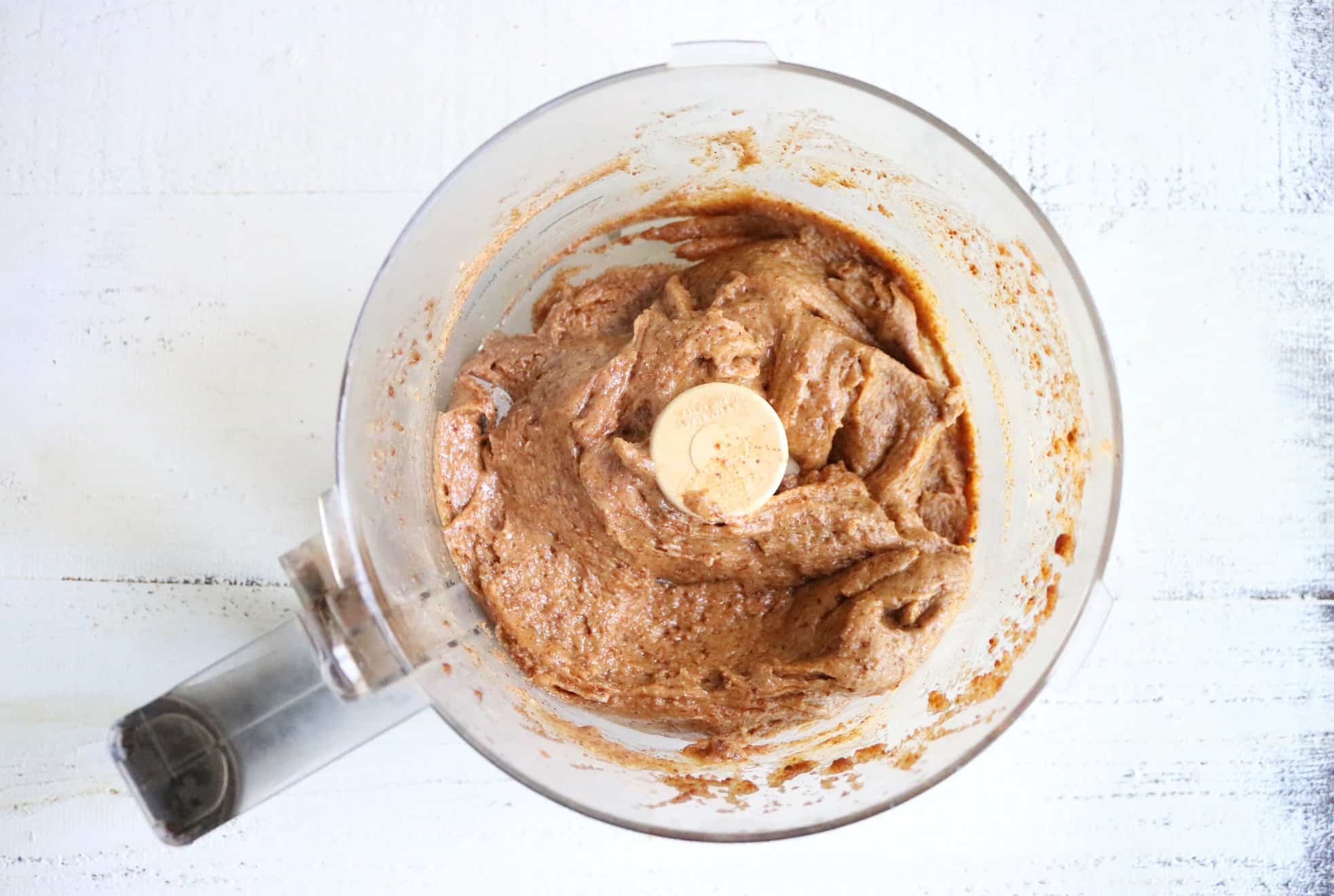 This is an overhead image of a food processor with an almond butter mixture in it. The food processor sits on a white surface.