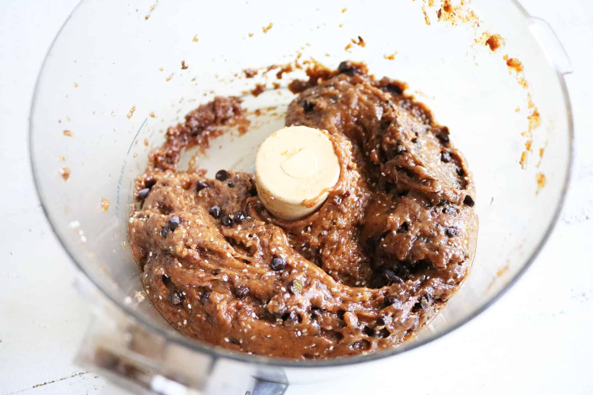 This image looks into a food processor with an almond butter mixture with chia seeds and mini chocolate chips. The food processor sits on a white surface.