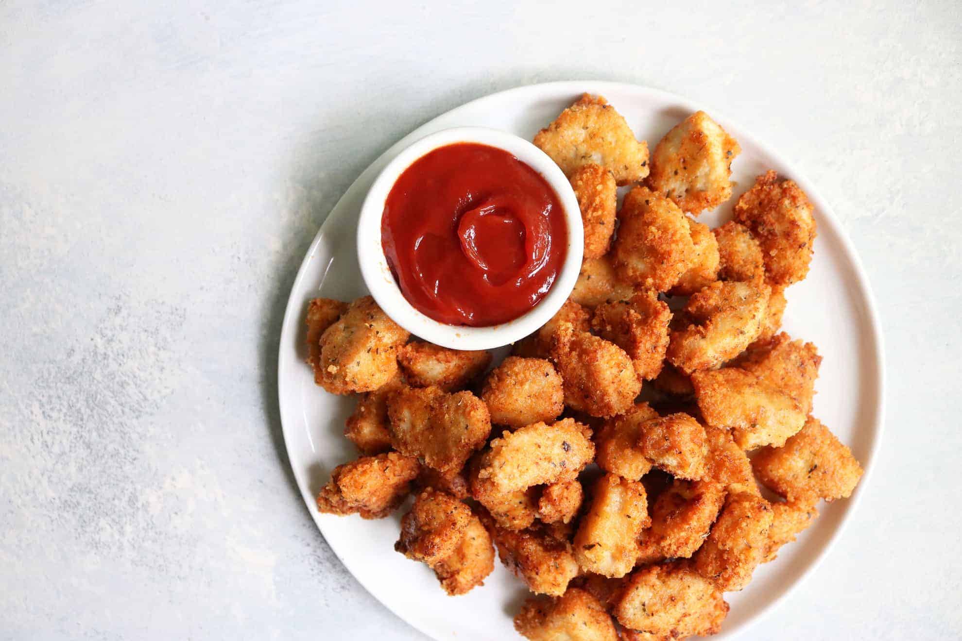 This is an overhead image of a white plate filled with chicken nuggets. A small white bowl filled with ketchup is also on the plate with the nuggets. The plate sites on a white and grey surface.