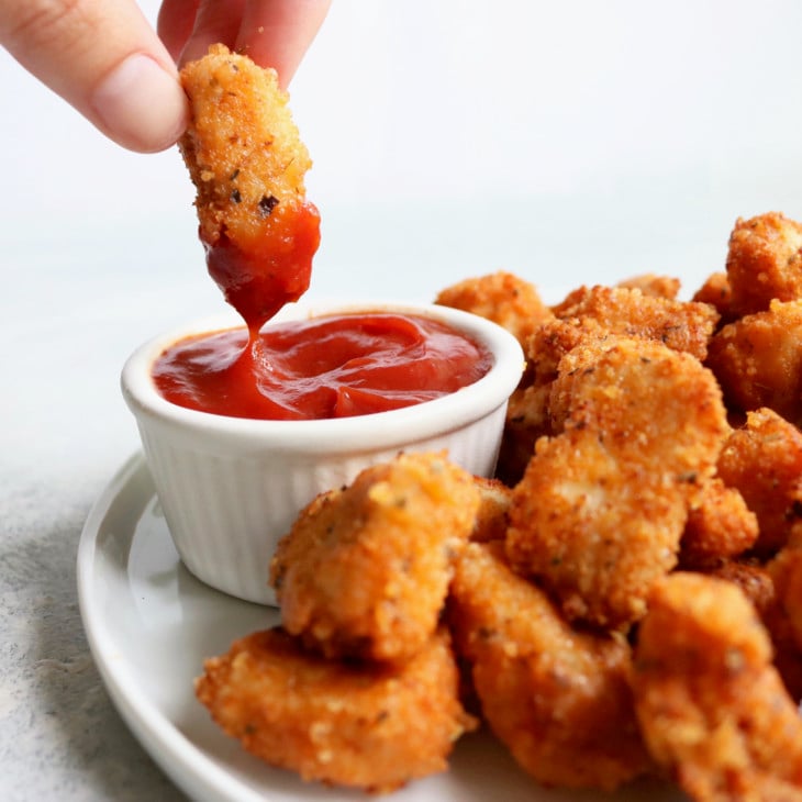 This is a side view of a white plate filled with chicken nuggets. A hand is holding one nugget above the plate and dipping it in a small white bowl of ketchup. The counter and background is white.