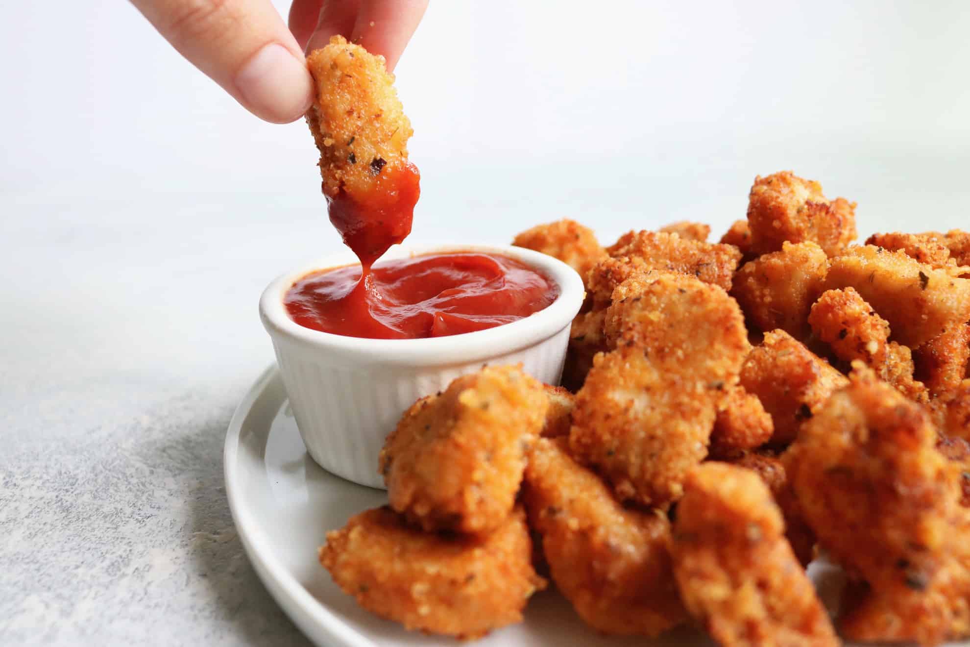 This is a side view of a white plate filled with chicken nuggets. A hand is holding one nugget above the plate and dipping it in a small white bowl of ketchup. The counter and background is white.