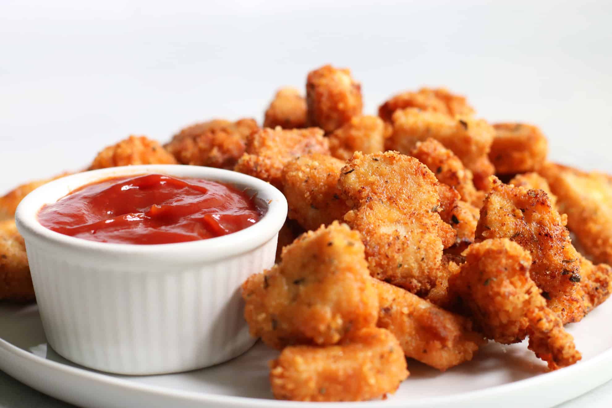 This is a side view of a white plate filled with chicken nuggets. A small white bowl filled with ketchup is also on the plate. The counter and background is white.