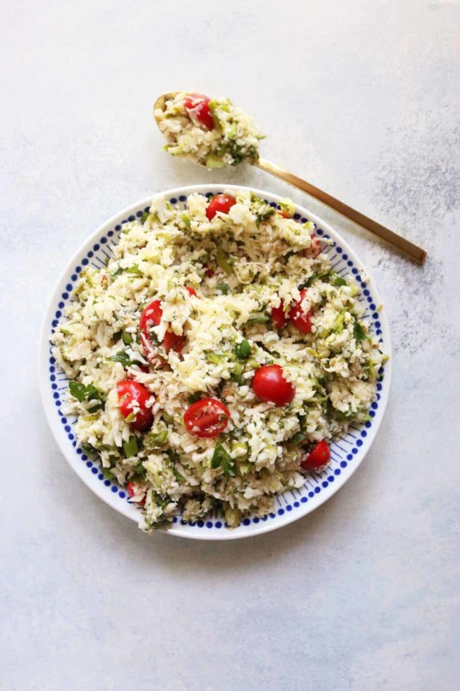 This is an overhead image of a blue and white bowl with cauliflower rice, sliced tomatoes, and herbs. The bowl sits on a white and grey counter with a gold spoon to the top of the bowl. 