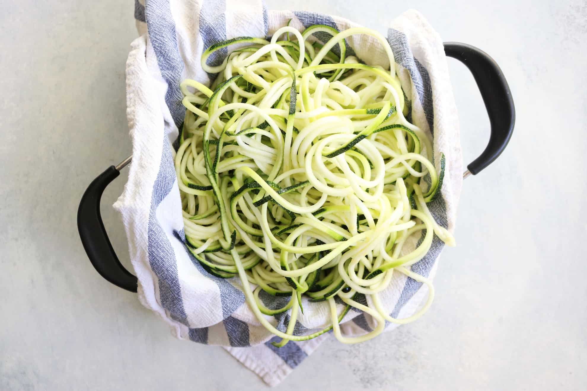 This is an overhead image of zoodles in a blue and white tea towel in a colander. The colander sits on a white counter.