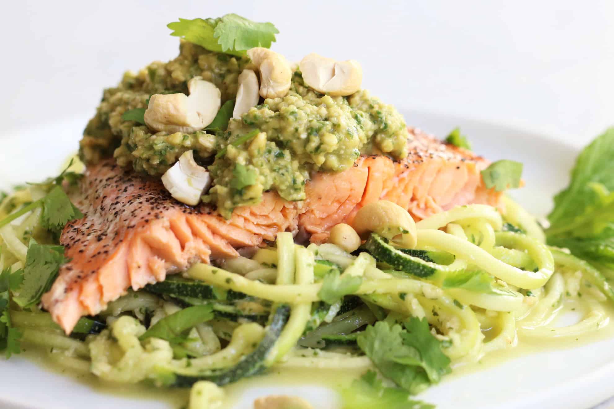 This is a side view of a white plate with zoodles, salmon and pesto on top. The dish is garnished with cashews and fresh cilantro leaves. 