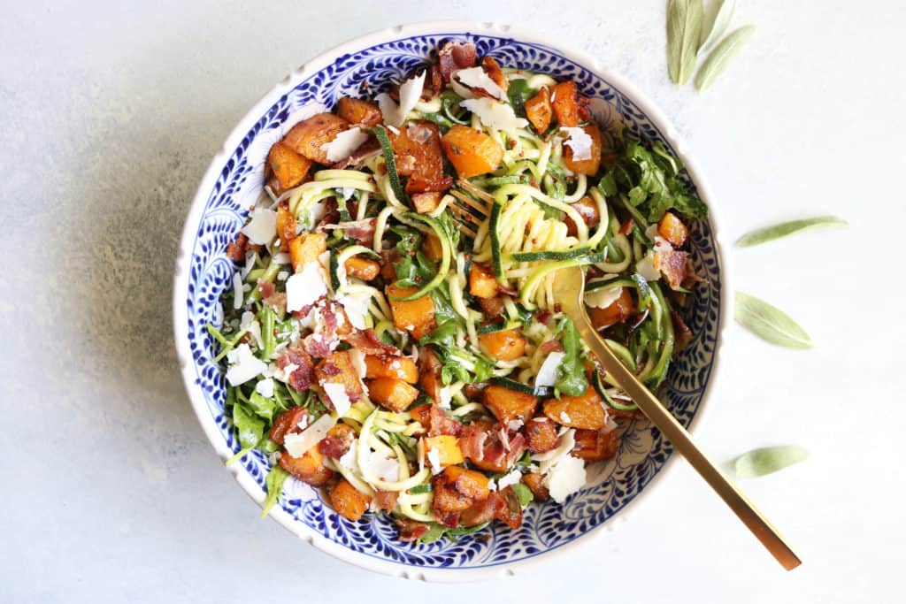 This is an overhead image of zoodle salad with butternut squash, bacon, and cheese. Fresh sage leaves are to the right of the bowl. A fork twirls the zoodles and is laying in the side of the bowl. 