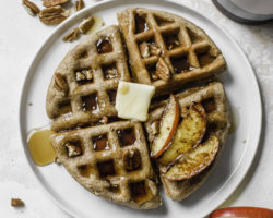 white plate with pumpkin waffles with butter, syrup, pecans, and sliced apples on top