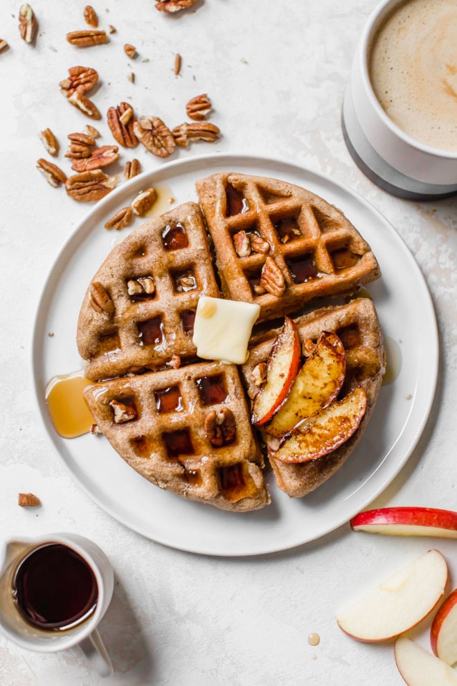 white plate with pumpkin waffles with butter, syrup, pecans, and sliced apples on top