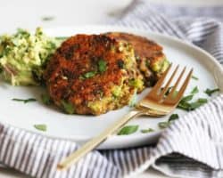 This is a side view of two Broccoli Cheddar Patties sitting on a white plate. There is some smashed avocado off to the side of the patties and they are garnished with chopped herbs. A gold fork lays next to the patties alongside the white plate. A grey pinstripe tea towel is underneath the plate.