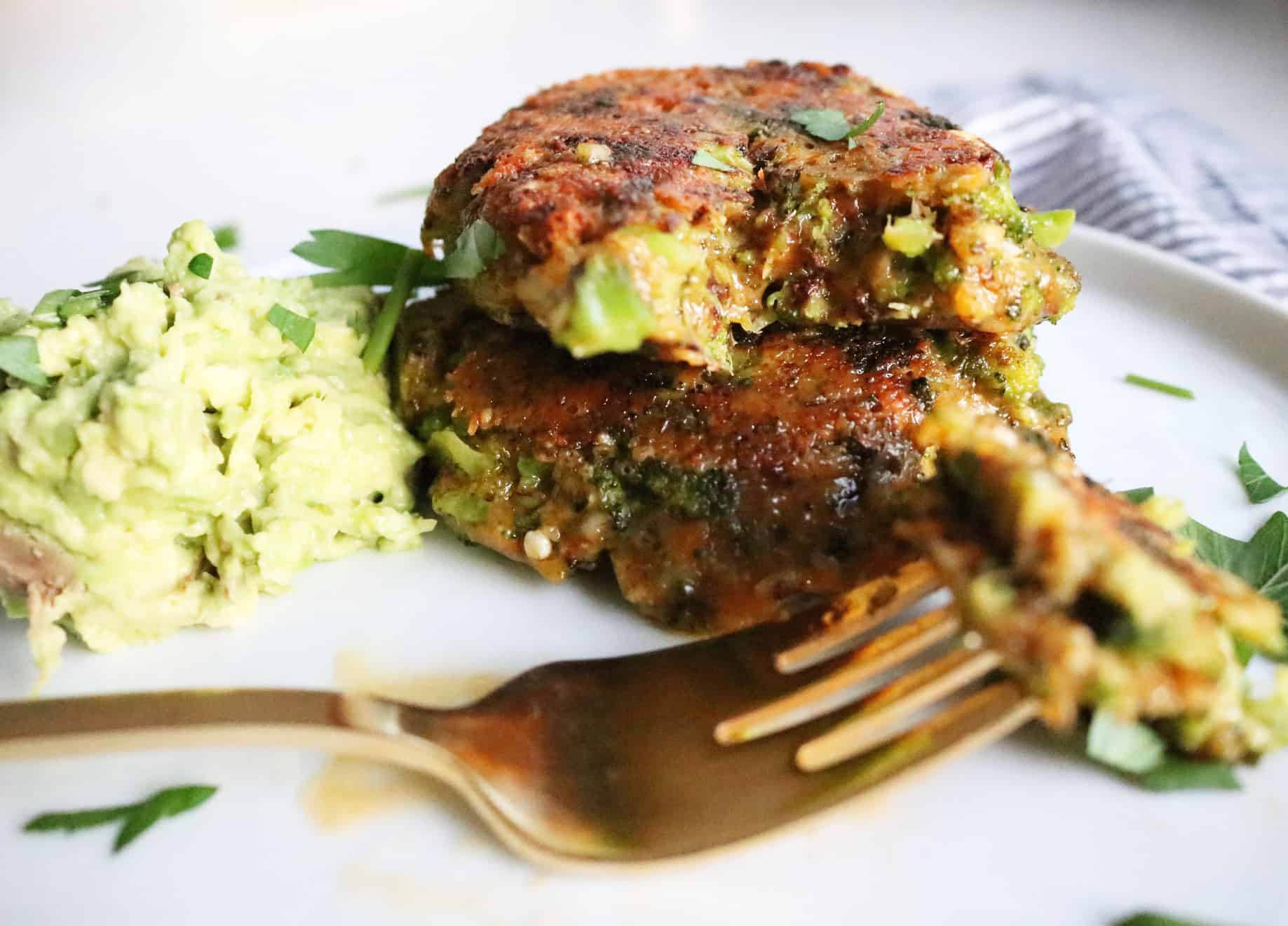 This is a side view of Broccoli Cheddar Patties. The two patties are stacked on top of each other and a bite is taken out of the top patty. A gold fork is blurred in the foreground with the bite of the broccoli patty. The patties sit on a white plate with smashed avocado off to the side. The plate sits on a white surface with a grey pinstripe tea towel underneath. 