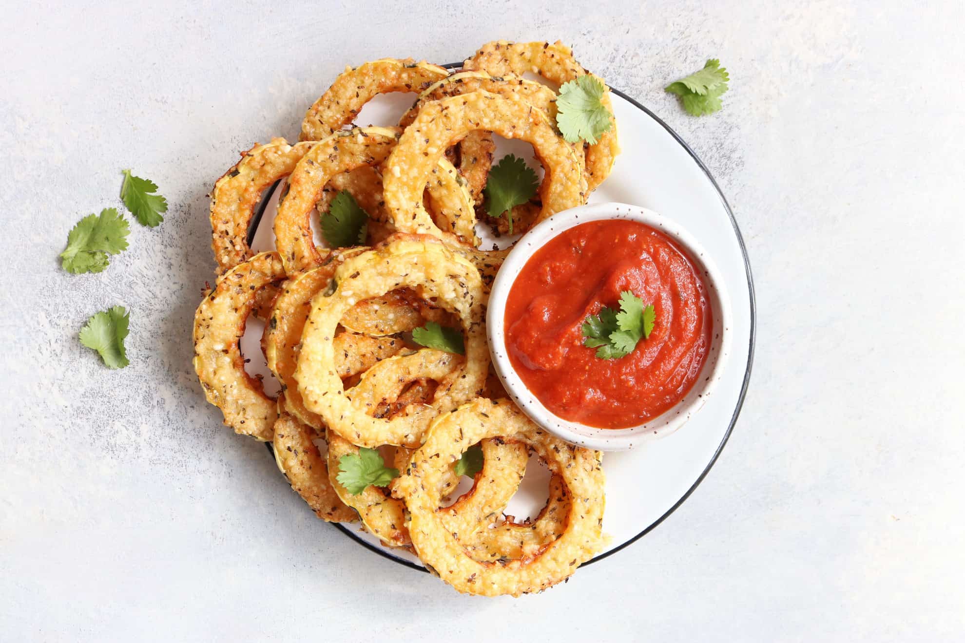 plate with Parmesan Crusted Delicata Squash rings with a small bowl of marinara sauce and some fresh herbs sprinkled on top on a white background