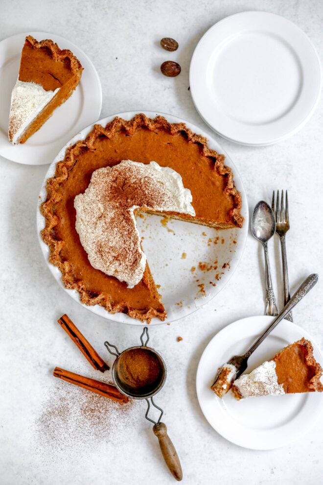 This is an overhead vertical image of a pumpkin pie. The pumpkin pie is in a white pie dish with a light brown pinched pie crust, a large dollop of whipped cream and a sprinkle of ground cinnamon. The pie dish sits on a light grey, almost white surface. Antique silverware is to the right side of the pie dish. To the top right corner of the image is a couple small white plates stacked on top of each other. A small white plate with a slice of pumpkin pie is to the bottom right corner and another plate with a slice of pumpkin pie is in the bottom right corner. A small handheld sifter with ground cinnamon is to the bottom center of the image with two cinnamon sticks.