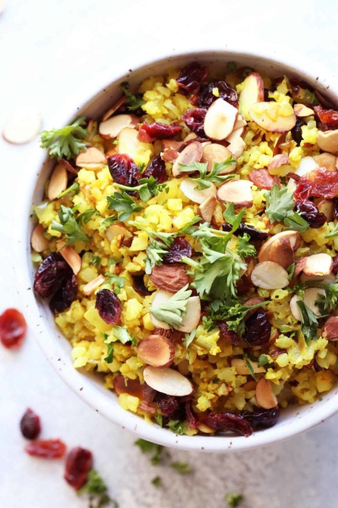 This is an overhead image of a bowl of yellow turmeric rice with slivered almonds, dried cranberries, and fresh herbs. The bowl sits on a white counter with more dried cranberries and herbs around it.
