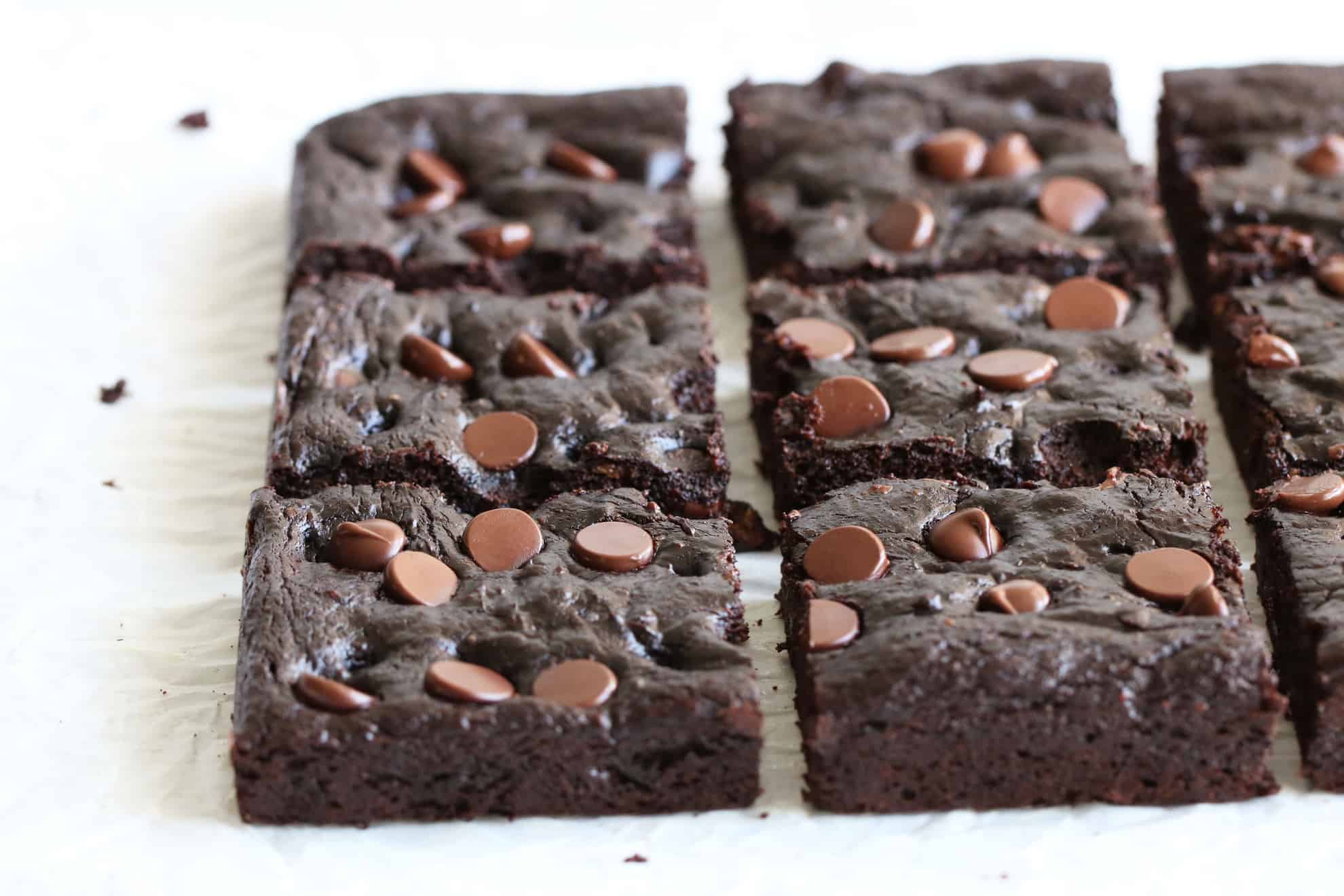 This is a side view of healthy avocado brownies with chocolate chips on the tops. The brownies are on a white surface and one brownie square has a bite taken out of it. 